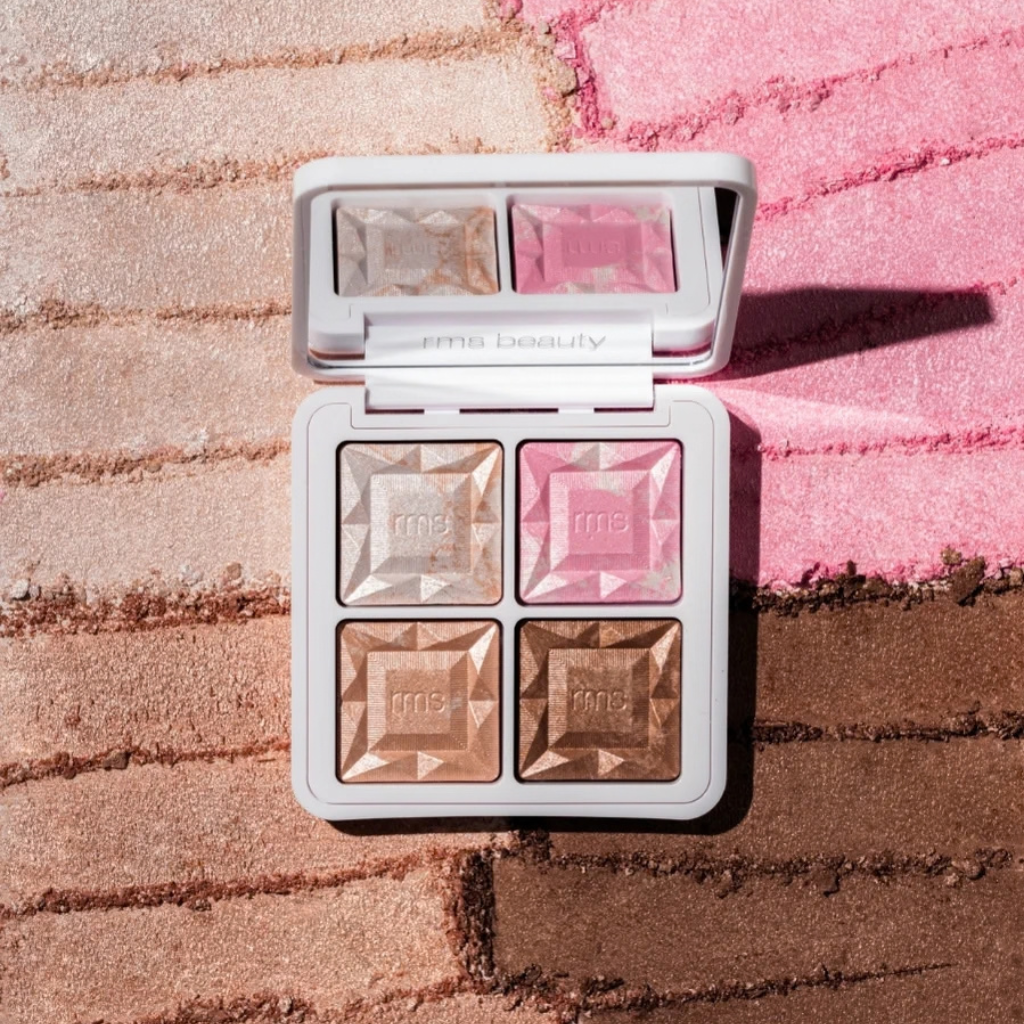 An open RMS Beauty four-pan highlighter palette with faceted pressed powder pans in champagne, pink, and bronze shades, set against a background of textured, colored powder in pink and brown.