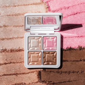 An open RMS Beauty four-pan highlighter palette with faceted pressed powder pans in champagne, pink, and bronze shades, set against a background of textured, colored powder in pink and brown.