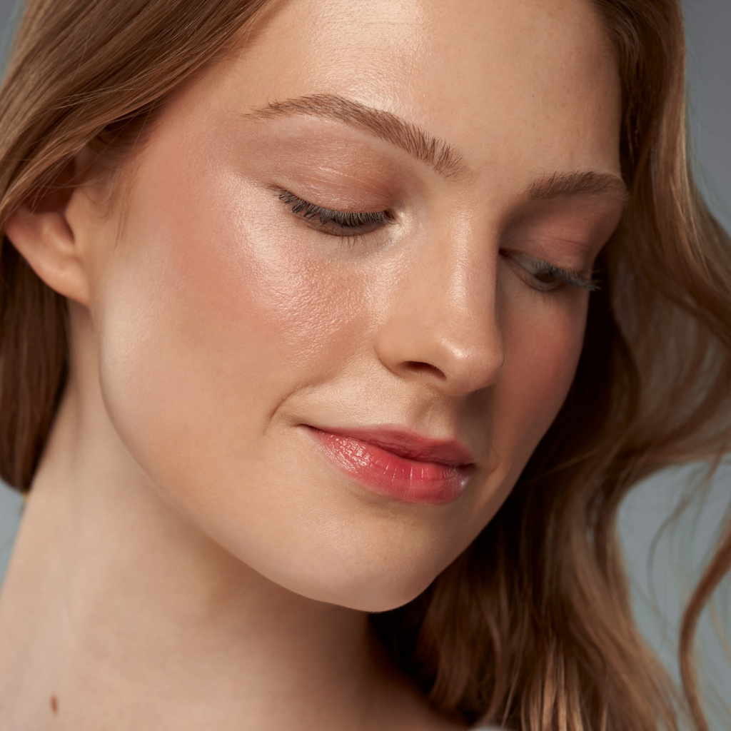 Close-up of a woman with closed eyes, wearing a glossy pink lip product and soft warm-toned makeup against a muted gray-blue background.