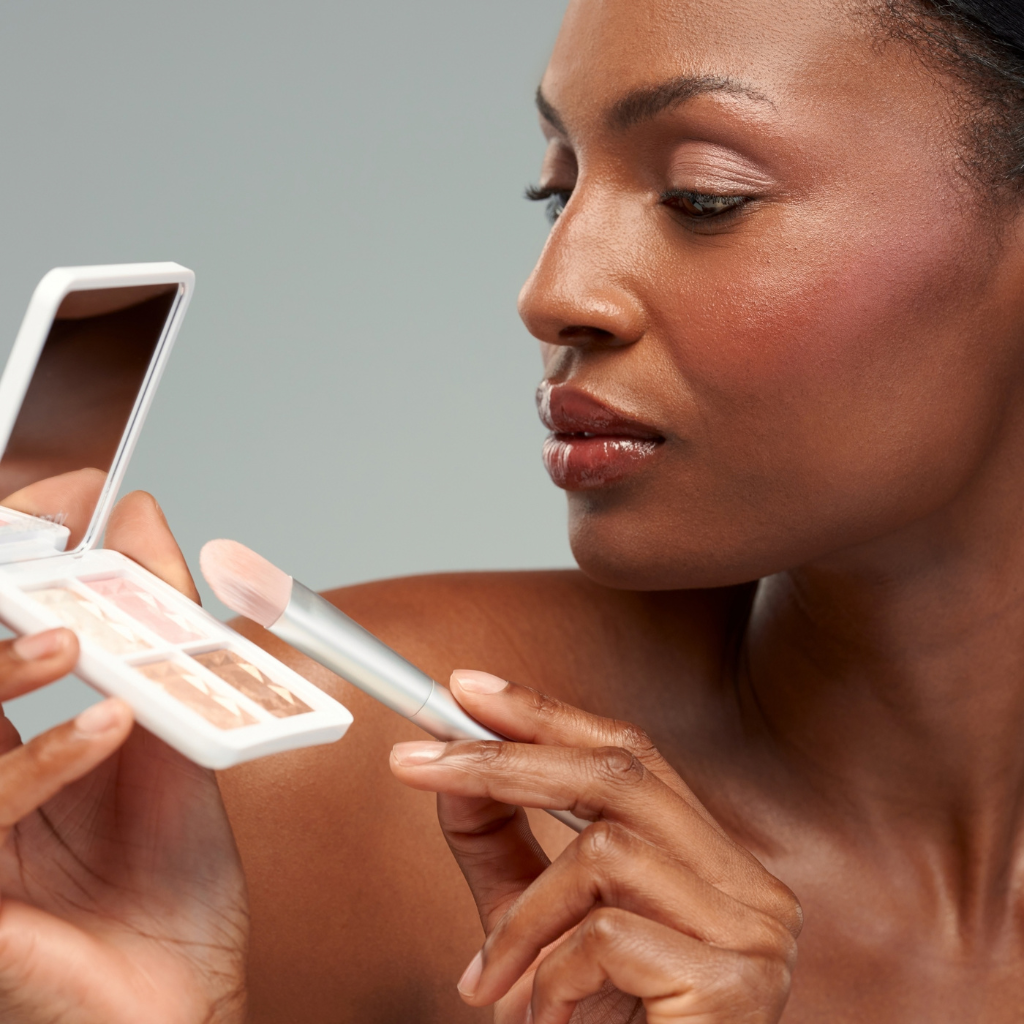 A close-up lifestyle shot of a woman applying makeup, holding a compact mirror and a makeup brush with a light pink brush tip against a solid gray background.