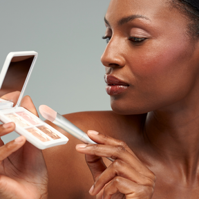 A close-up lifestyle shot of a woman applying makeup, holding a compact mirror and a makeup brush with a light pink brush tip against a solid gray background.